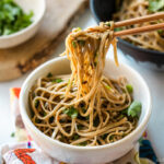 Chopsticks pulling noodles from a white bowl containing Soba Noodle Salad topped with sesame seeds and chopped cilantro and green onions.