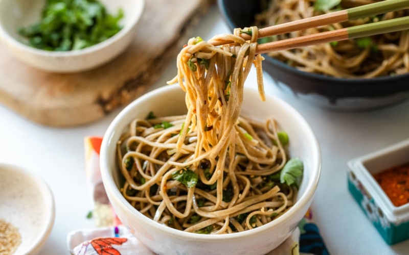 Chopsticks pulling noodles from a white bowl containing Soba Noodle Salad topped with sesame seeds and chopped cilantro and green onions.