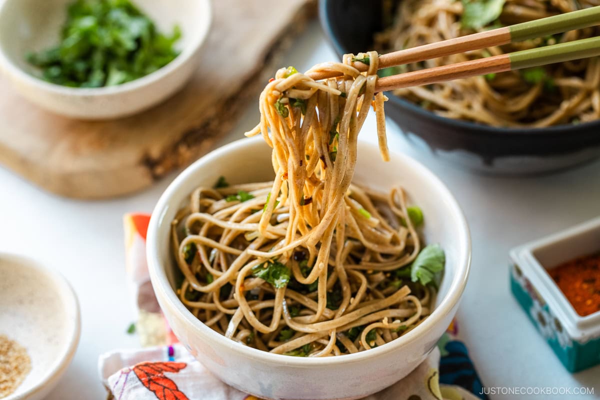 Chopsticks pulling noodles from a white bowl containing Soba Noodle Salad topped with sesame seeds and chopped cilantro and green onions.