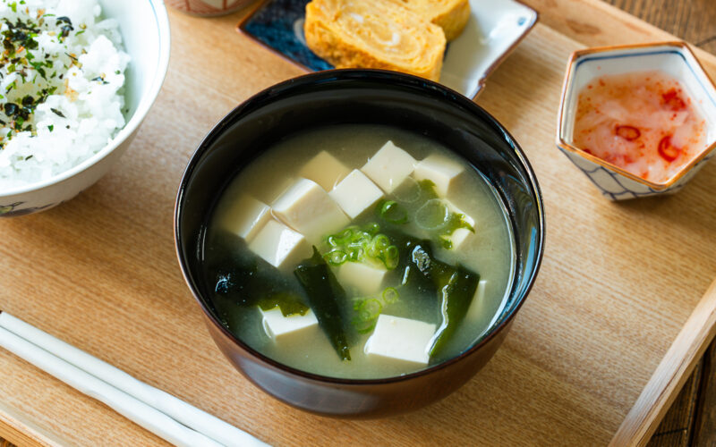 A light-colored wooden tray containing a bowl of miso soup with soft tofu, wakame seaweed, and thinly sliced green onions, along with a bowl of steamed rice, a small plate of tamagoyaki, and a smalll side dish.