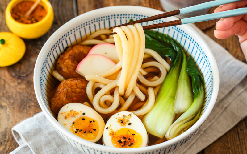 A large blue and white bowl containing udon noodle soup topped with fish cake, bok choy, fish cakes, and jammy soft boiled eggs in a savory dashi broth.