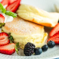 A white plate containing Japanese souffle pancakes dusted with powdered sugar, fresh mixed berries, and whipped cream.