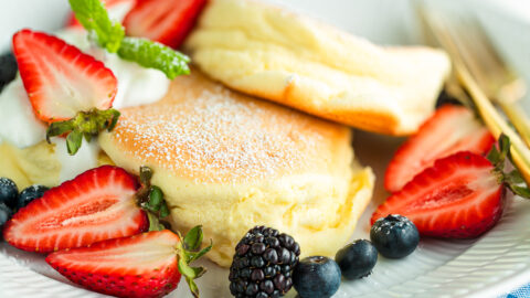 A white plate containing Japanese souffle pancakes dusted with powdered sugar, fresh mixed berries, and whipped cream.
