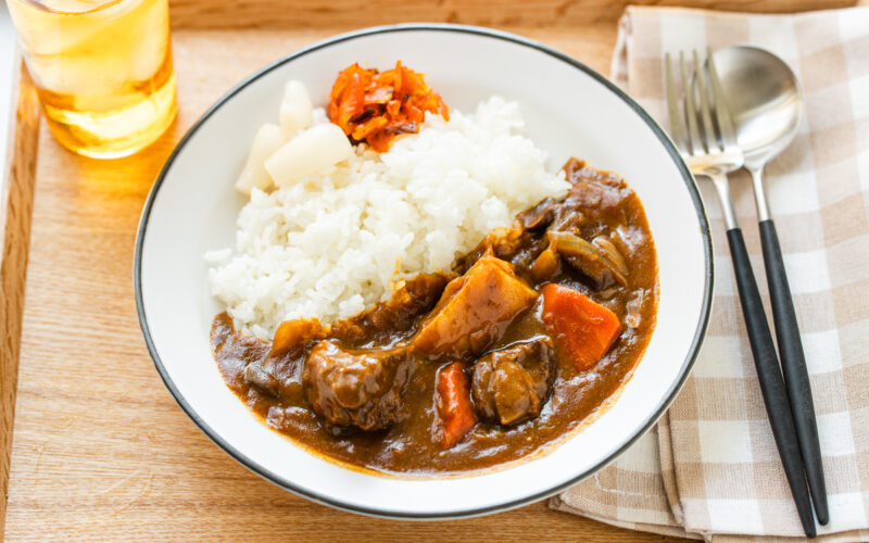 A round white plate containing Japanese beef curry consisting of tender beef, potatoes, carrots, mushrooms, and curry roux, served over steamed rice.