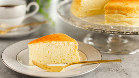 A white and gray rim plate containing a slice of Japanese souffle cheesecake. A cake stand behind containing the whole cake.