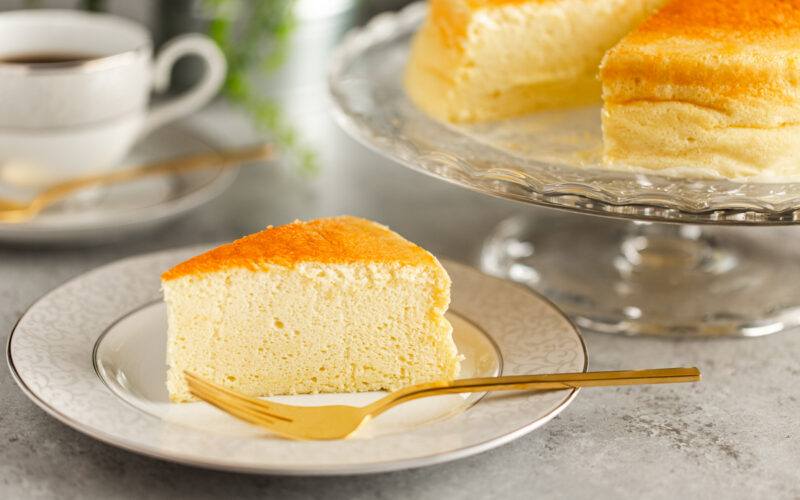 A white and gray rim plate containing a slice of Japanese souffle cheesecake. A cake stand behind containing the whole cake.
