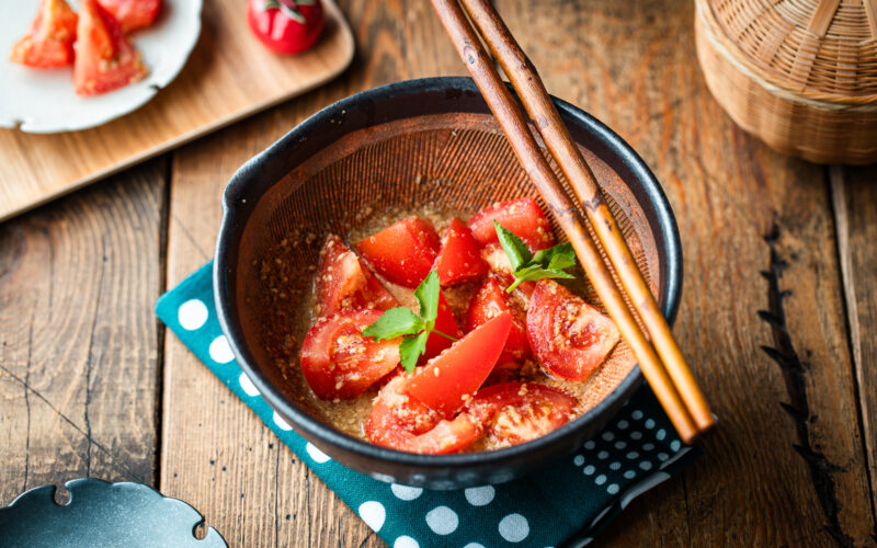 A Japanese black mortar containing cut tomato wedges dressed in ground sesame ponzu seasoning, garnished with mitsuba leaves on top.