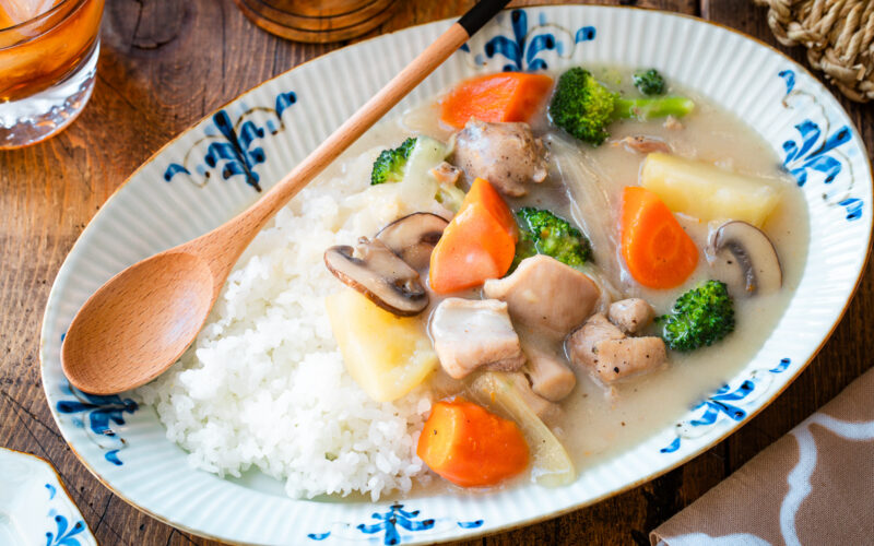 An oval-shaped plate holds a comforting serving of Japanese cream stew, featuring tender chicken, carrots, potatoes, mushrooms, and broccoli in a rich, creamy white sauce. The stew is generously ladled over a bed of fluffy steamed rice, creating a hearty and satisfying meal.