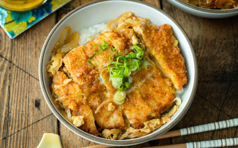 A white bowl containing Katsudon, pork cutlet rice bowl, where golden brown tonkatsu, drizzled egg, and tender onion are simmered in a savory dashi broth.