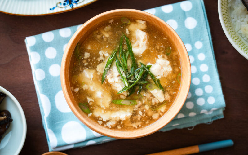 Wooden bowls filled with Pork Tofu Soup, served alongside steamed rice and a chicken dish on the table.