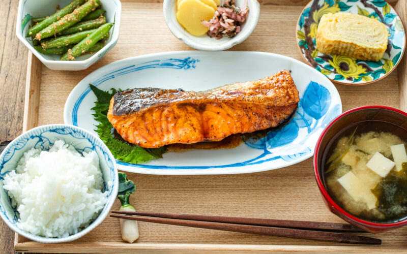 A Japanese meal consisting of small dishes or bowls, each plating Teriyaki Salmon, steamed rice, miso soup, pickles, tamagoyaki, and green bean gomaae.