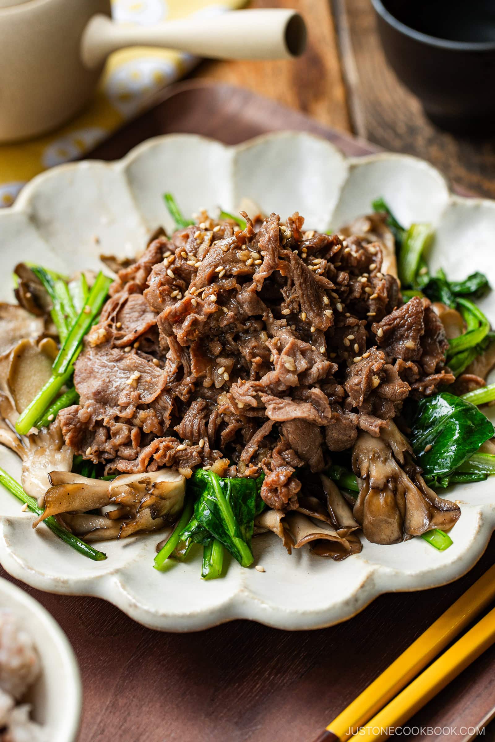 A white scalloped plate containing stir-fried Butter Ponzu beef with komatsuna green and maitake mushrooms.