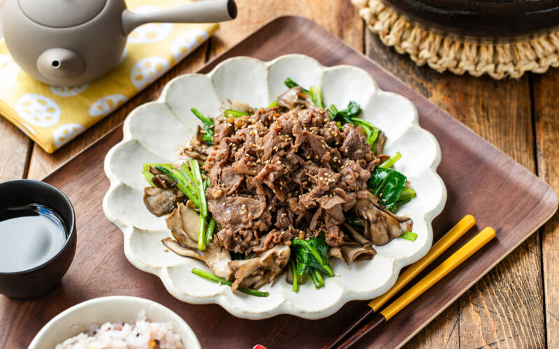 A white scalloped plate containing stir-fried Butter Ponzu beef with komatsuna green and maitake mushrooms.