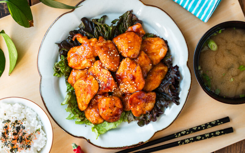 A white plate containing Honey Gochujang Chicken sprinkled with sesame chicken served on green salad.