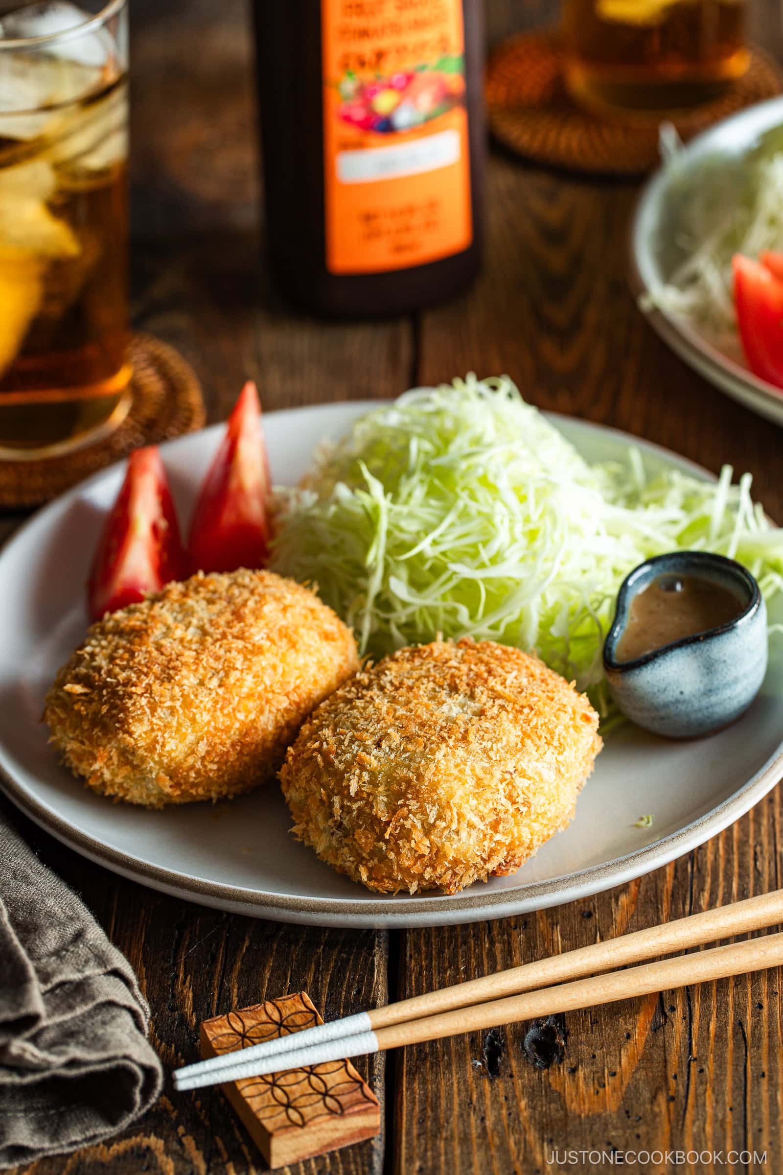 A round white plate containing Japanese croquettes, shredded cabbage, tomato wedges, and a tiny container with sesame dressing.