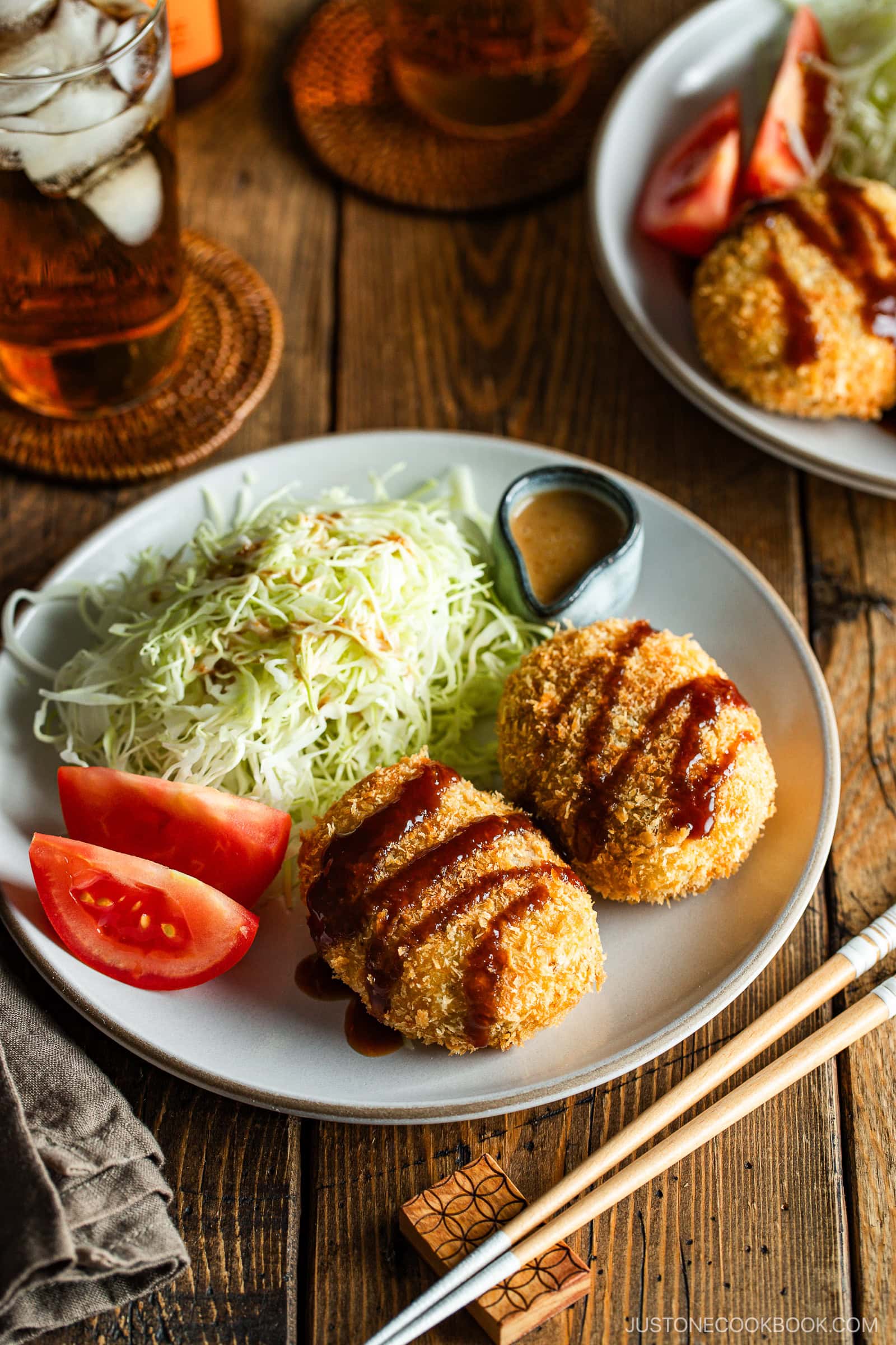 A round white plate containing Japanese croquettes, shredded cabbage, tomato wedges, and a tiny container with sesame dressing.