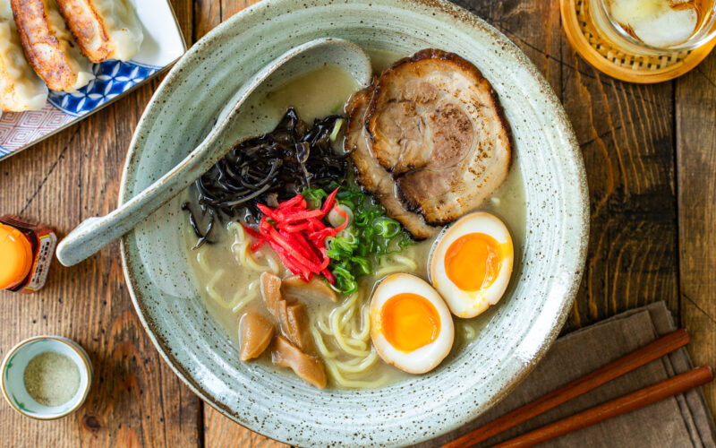 A rustic bowl containing noodles with tonkotsu broth, topped with chashu, ramen eggs, menma, kikurage, ginger, and chopped green onion. On the table, a plate of crispy gyoza and a glass of mugicha are served together.
