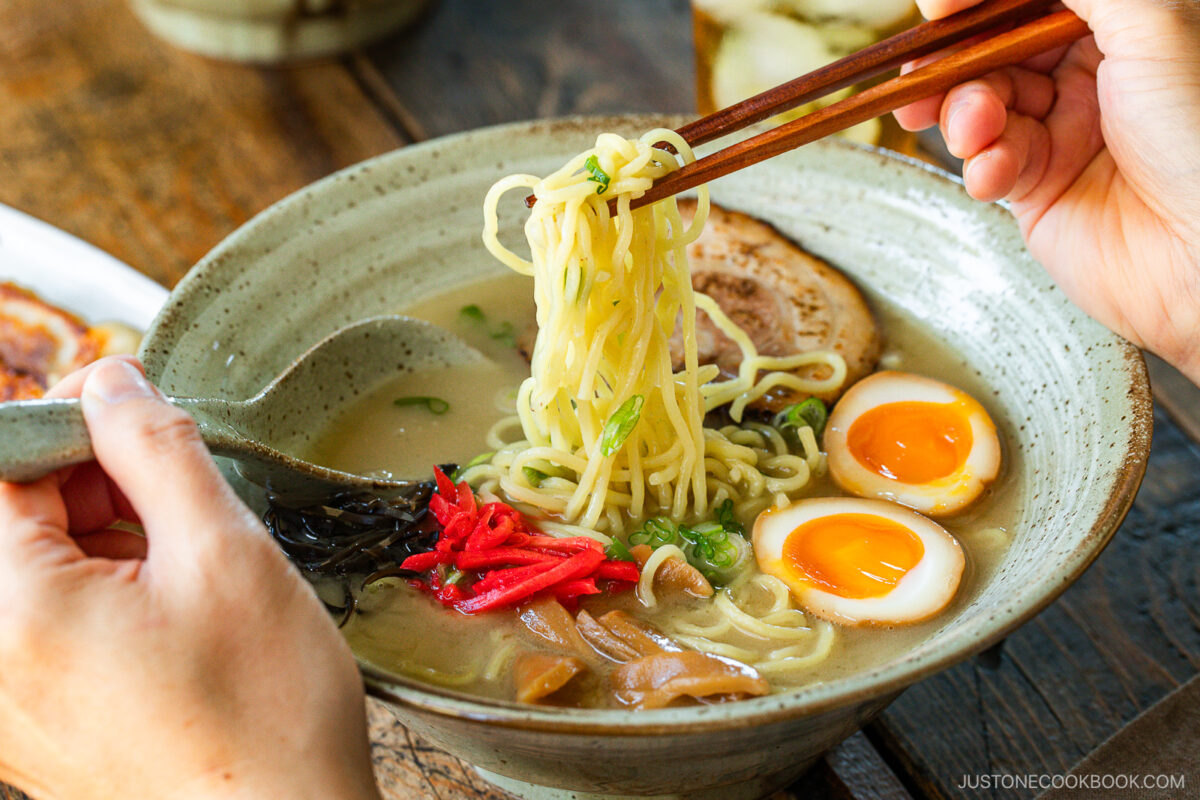 A rustic bowl containing noodles with tonkotsu broth, topped with chashu, ramen eggs, menma, kikurage, ginger, and chopped green onion. On the table, a plate of crispy gyoza and a glass of mugicha are served together.