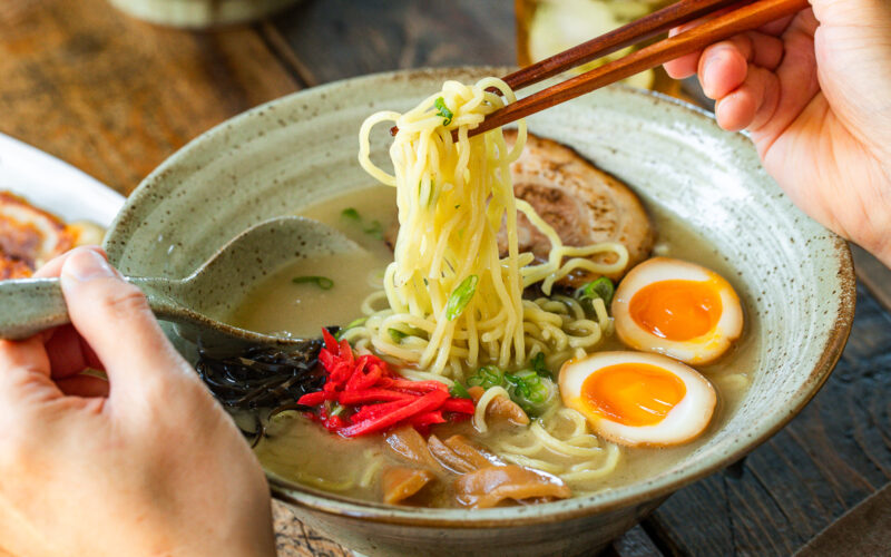 A rustic bowl containing noodles with tonkotsu broth, topped with chashu, ramen eggs, menma, kikurage, ginger, and chopped green onion. On the table, a plate of crispy gyoza and a glass of mugicha are served together.