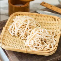 Two bundles of freshly made, uncooked udon noodles rest on a bamboo tray, placed atop a wooden cutting board with a ruler, rolling pin, and kitchen knife in the background.