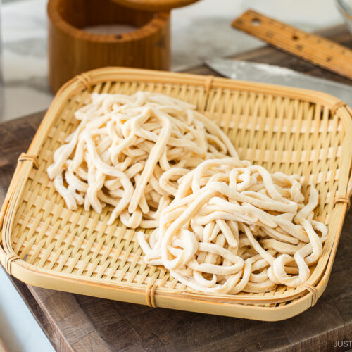 Two bundles of freshly made, uncooked udon noodles rest on a bamboo tray, placed atop a wooden cutting board with a ruler, rolling pin, and kitchen knife in the background.