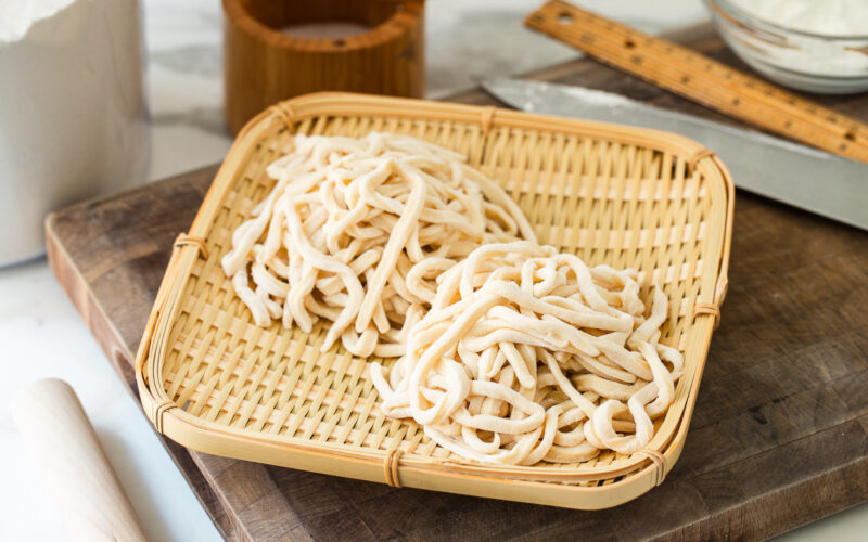 Two bundles of freshly made, uncooked udon noodles rest on a bamboo tray, placed atop a wooden cutting board with a ruler, rolling pin, and kitchen knife in the background.