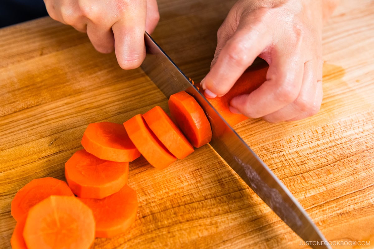 A person slicing a carrot into round pieces on a wooden cutting board with a large knife.