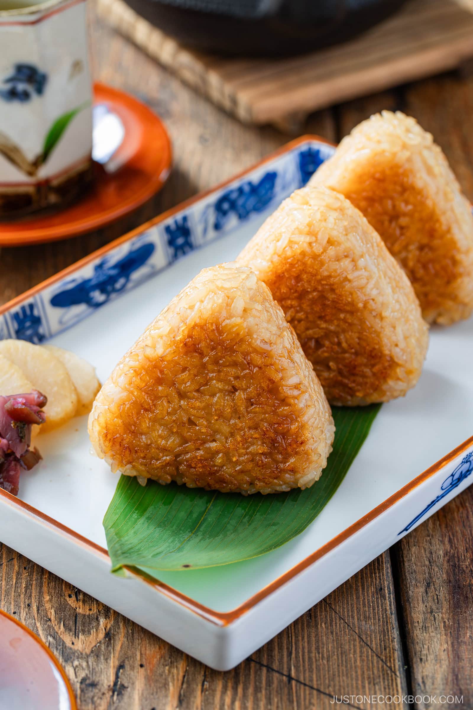 Three grilled triangular rice balls (yaki onigiri) with a golden-brown crust are arranged on a green leaf in a rectangular plate, accompanied by pickled vegetables on the side. The setting is on a wooden table.