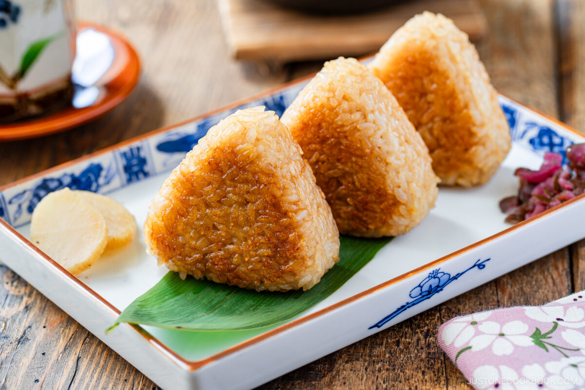 Three grilled Japanese rice balls (yaki onigiri) are arranged on a rectangular plate with a bamboo leaf, accompanied by pickled vegetables and a slice of pickled radish.