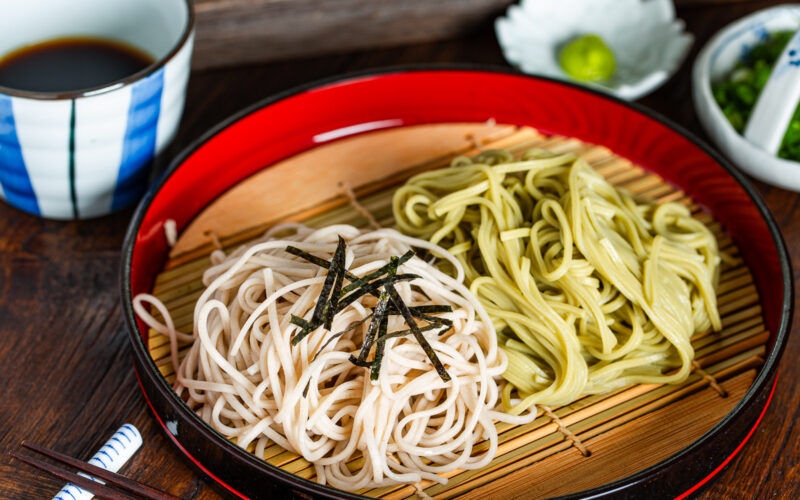 A red tray with bamboo mat holding two types of cold noodles, one white and one green, garnished with shredded seaweed, served with dipping sauce and condiments in bowls nearby. Chopsticks rest on the side.