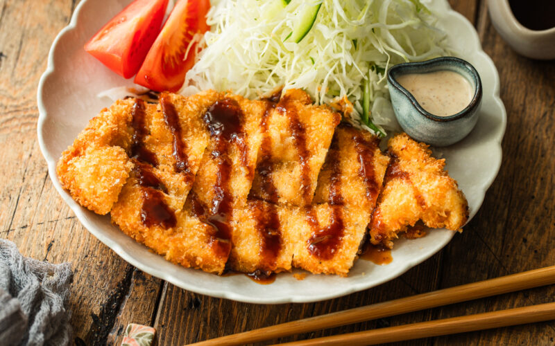 A plate of crispy breaded chicken cutlets drizzled with sauce, served with shredded cabbage, tomato wedges, chopsticks, and a small dish of creamy dressing on a wooden table.