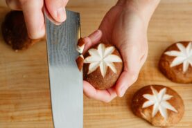 A person uses a knife to carve a decorative star pattern into the cap of a shiitake mushroom on a wooden cutting board.