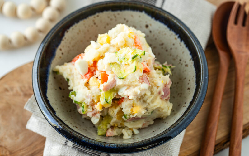 A bowl of Japanese potato salad with visible chunks of potato, ham, boiled egg, cucumber, and carrot, placed on a rustic plate beside wooden utensils and a beaded decoration.