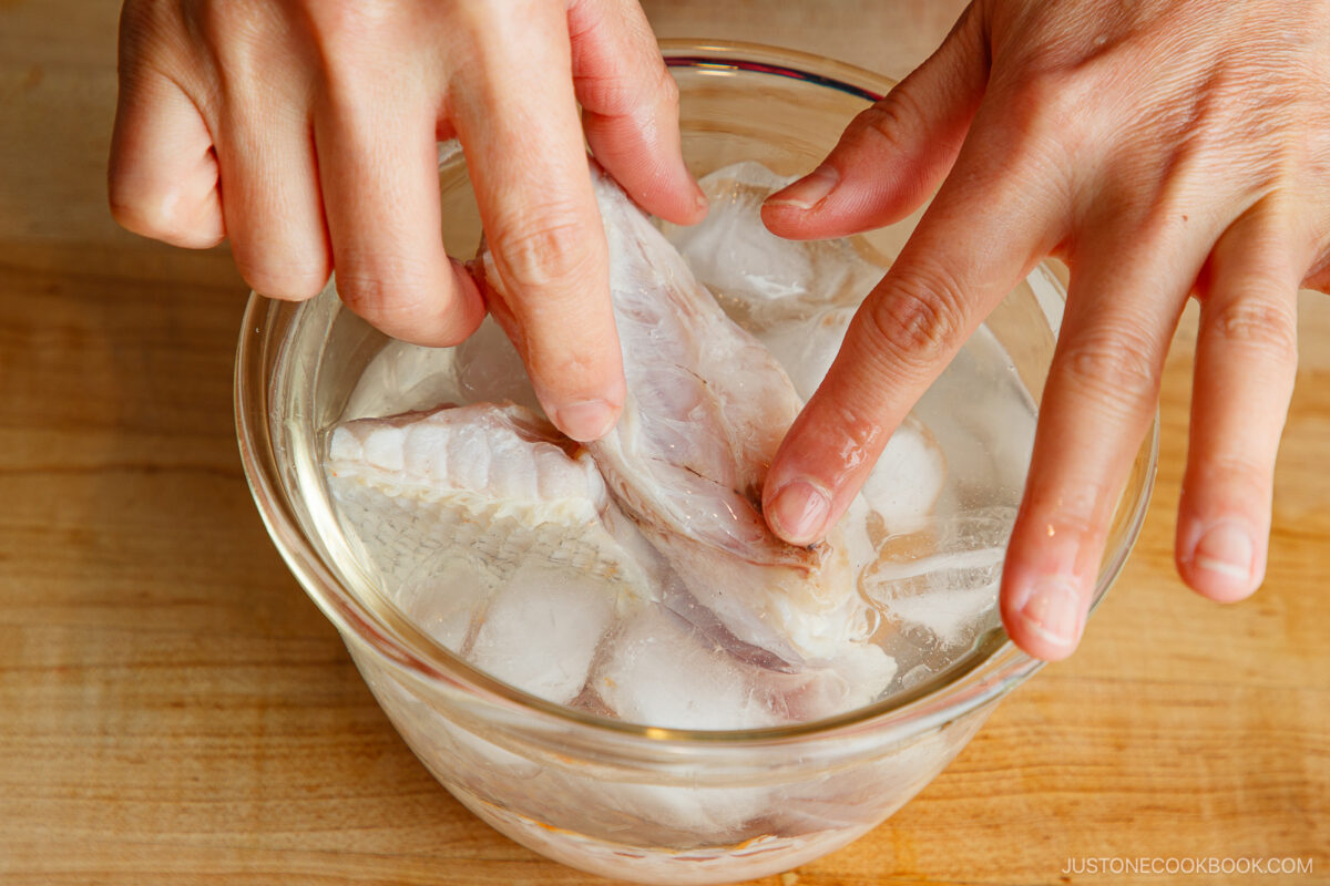 A person’s hands submerge raw fish pieces in a glass bowl filled with ice water on a wooden surface.