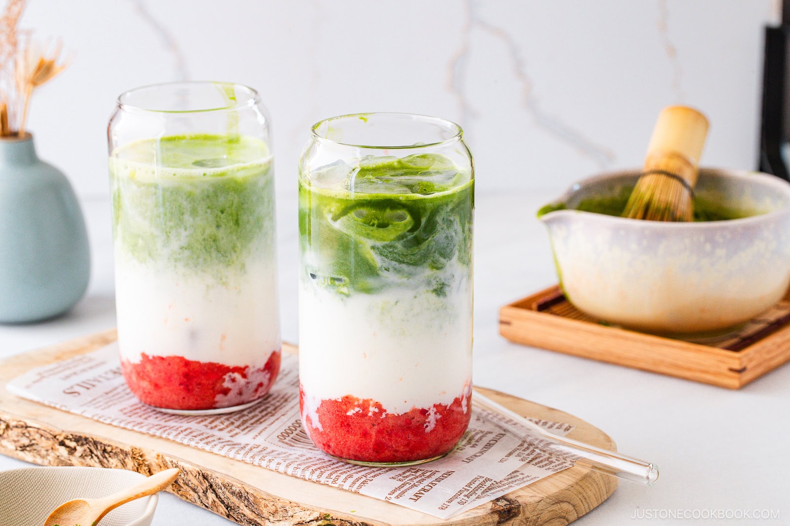 Two glasses filled with a layered drink: red strawberry puree at the bottom, white milk in the middle, and green matcha tea on top, served with ice on a wooden tray. A matcha whisk and bowl are in the background.
