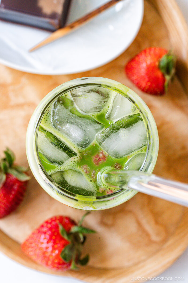 A top view of a glass filled with iced green matcha tea, surrounded by fresh strawberries on a wooden tray. A plate with a dark chocolate square is partially visible in the background.
