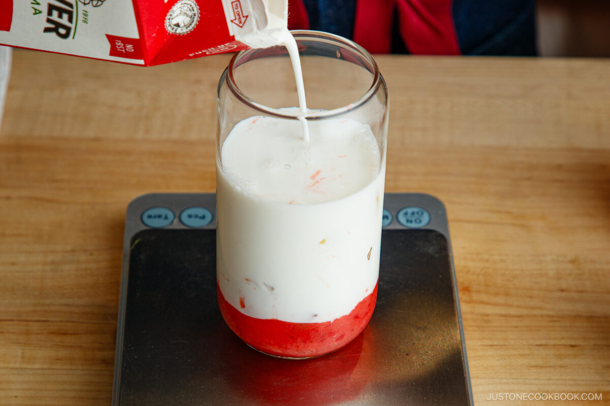 A glass with strawberry puree at the bottom is placed on a digital kitchen scale while milk is being poured in from a carton.