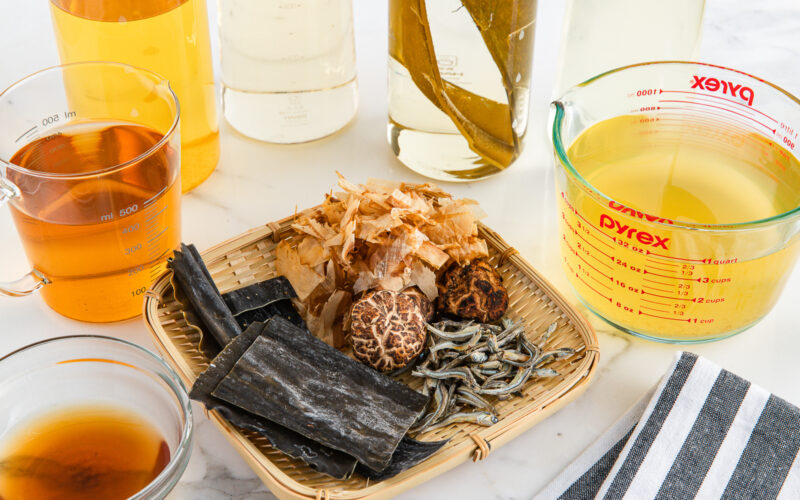 A bamboo tray with dried kombu, shiitake mushrooms, small fish, and bonito flakes sits on a white surface, surrounded by measuring cups and bottles filled with golden and clear broths. A striped cloth is nearby.