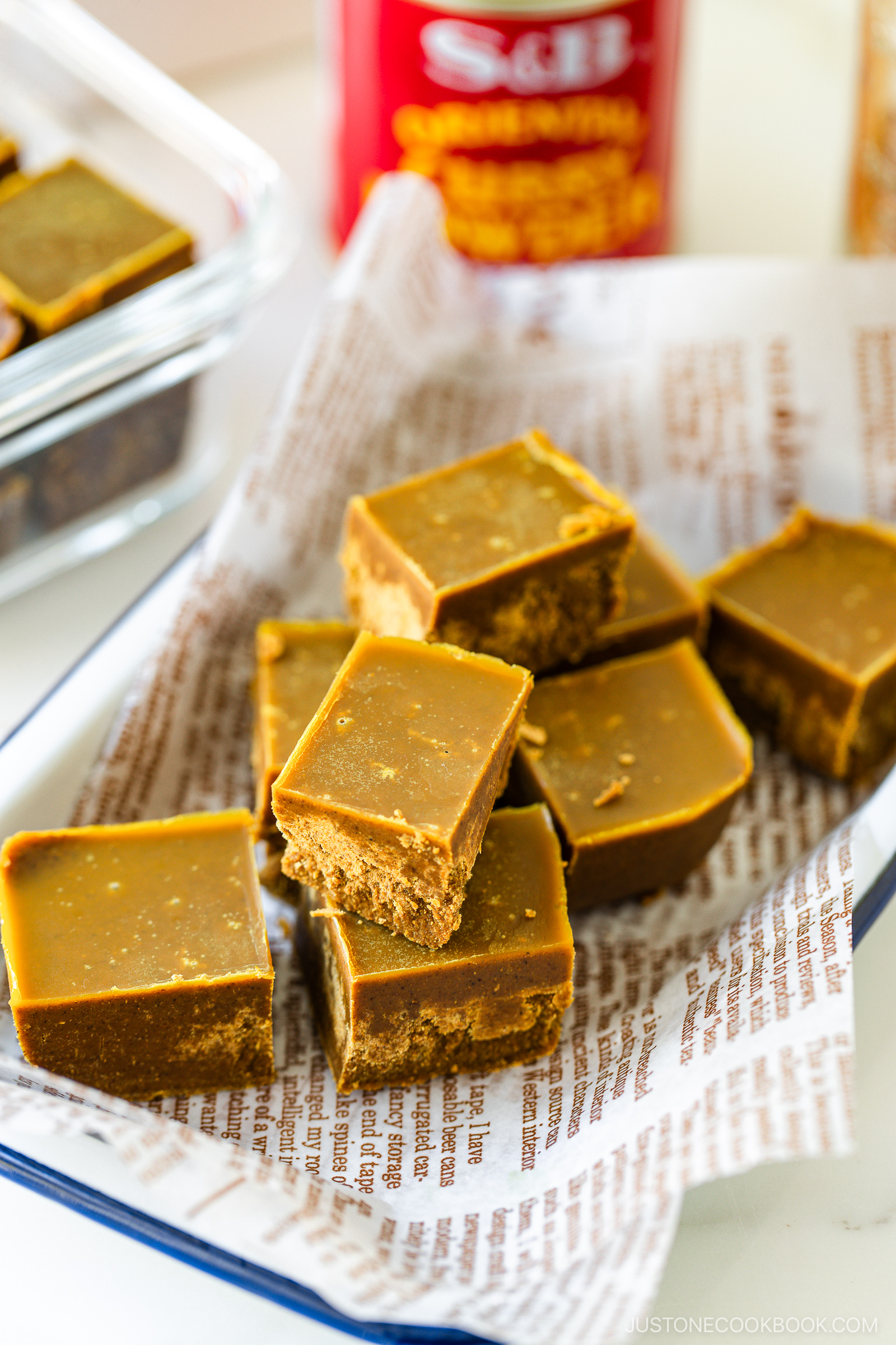 Several pieces of square-shaped brown fudge are stacked on a tray lined with parchment paper printed with text. More fudge and a red canister are visible in the background.