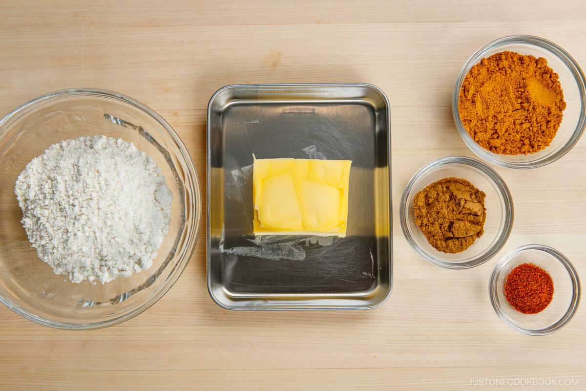 A metal tray with butter, a glass bowl of flour, and three small bowls containing brown spices are arranged on a light wooden surface.