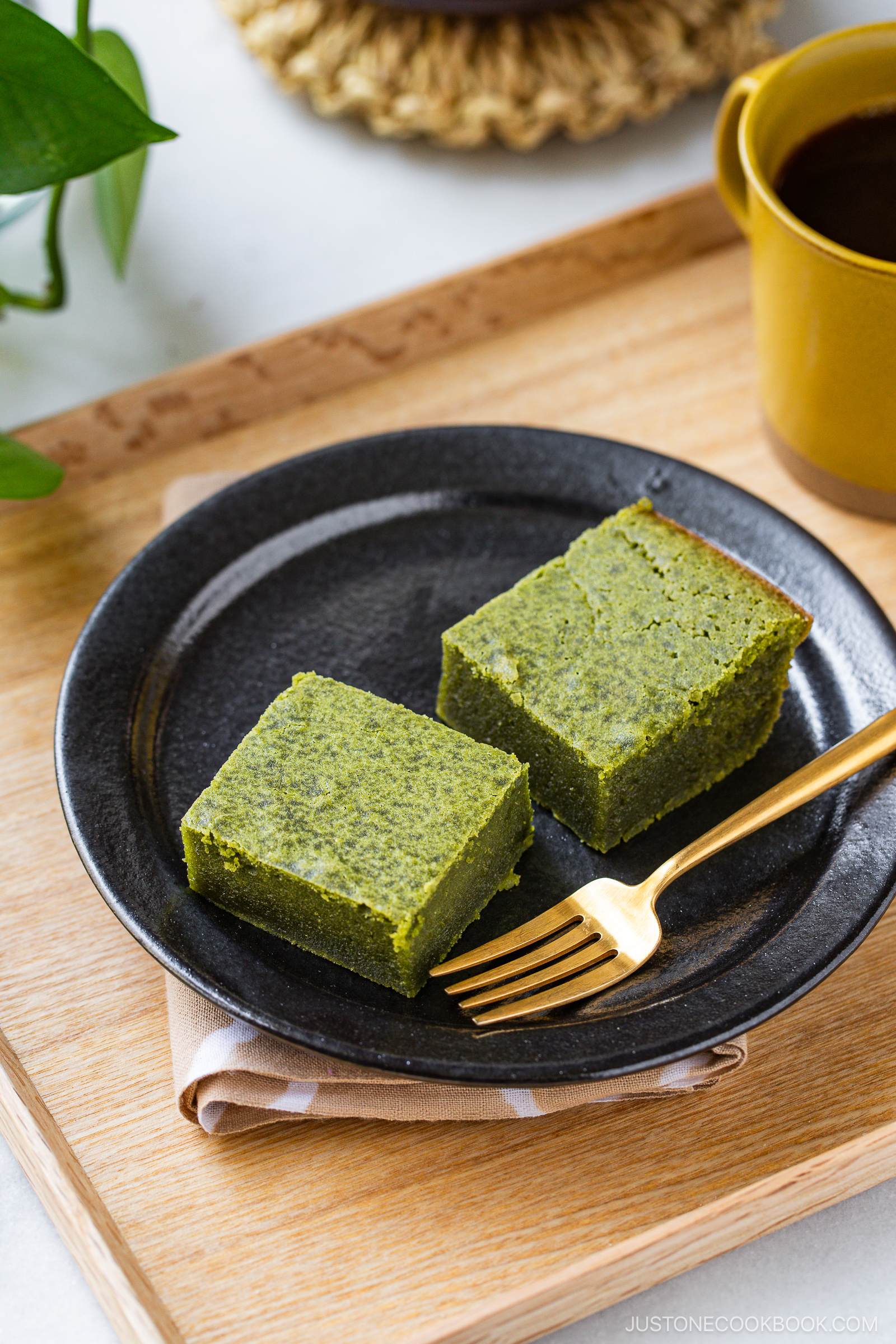 A black plate with two square slices of matcha cake and a gold fork, set on a wooden tray next to a yellow cup of coffee. A folded napkin is under the plate and a green plant is in the background.