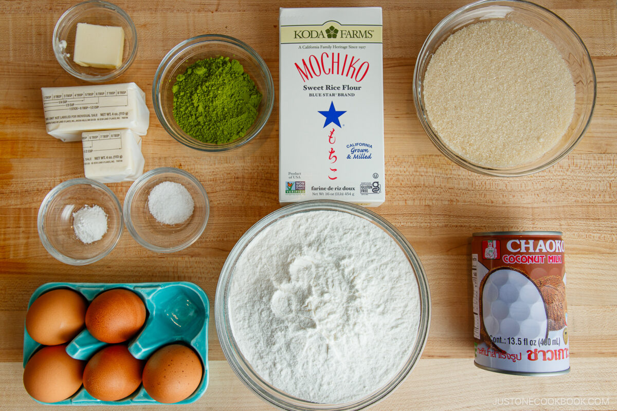 A top-down view of baking ingredients on a wooden surface, including eggs, butter, coconut milk, sugar, matcha powder, mochiko sweet rice flour, baking powder, and a bowl of flour.