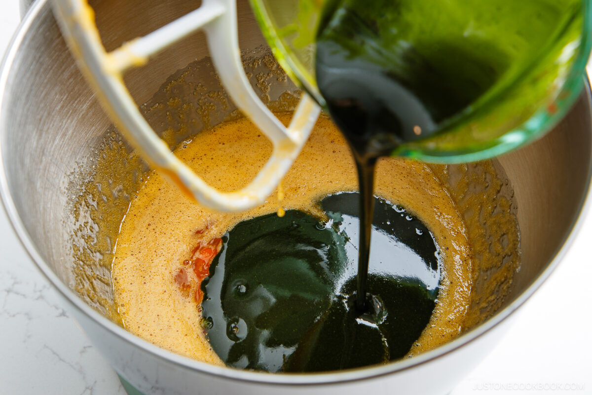 A close-up of thick dark molasses being poured from a green bowl into a metal mixing bowl with a yellow batter, with a white paddle attachment visible.