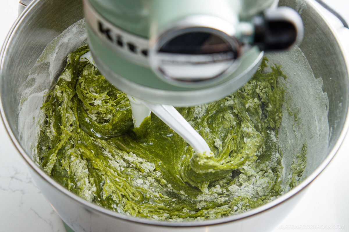 A stand mixer blends a green batter, likely matcha-flavored, in a metal mixing bowl. The paddle attachment is partially covered in the thick, vibrant mixture.