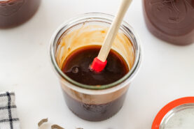 A glass jar filled with thick brown sauce, stirred with a small red and white spatula, sits on a white surface beside a jar lid, a striped cloth, and another jar.