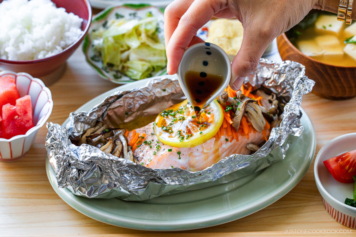 A hand pours sauce over baked salmon with lemon, mushrooms, and veggies in foil. Surrounding dishes include rice, salad, soup, watermelon, and tomato on a wooden table.