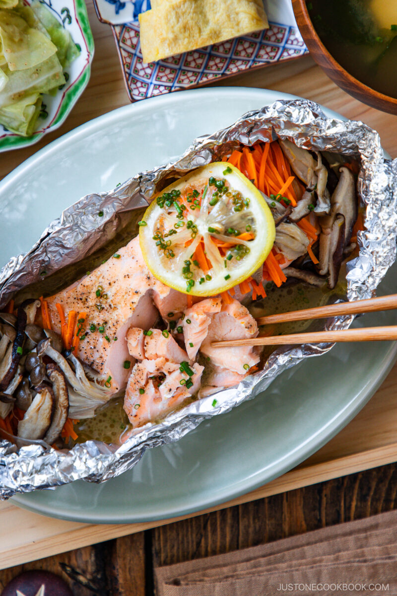 A foil packet with cooked salmon, sliced mushrooms, and julienned carrots, topped with a lemon slice and chopped herbs, on a plate. Chopsticks hold a piece of salmon. Other dishes are partially visible in the background.