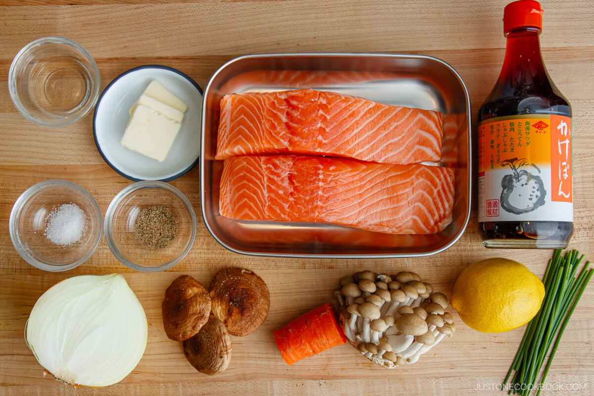 Salmon fillets in a tray with soy sauce, lemon, carrot, onion, mushrooms, chives, butter, salt, and pepper arranged on a wooden surface, viewed from above.
