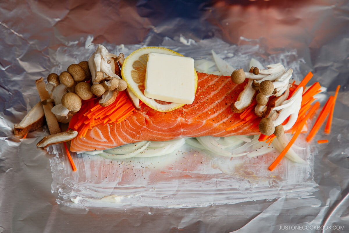Raw salmon fillet on a sheet of foil topped with a slice of lemon, a pat of butter, enoki and shimeji mushrooms, surrounded by sliced onions and julienned carrots, ready for cooking.