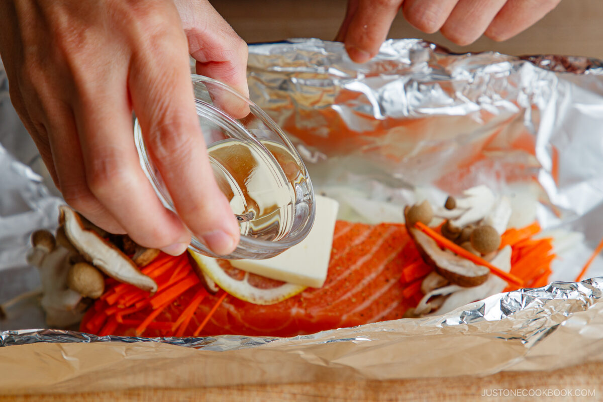 Close-up of hands pouring liquid from a small glass cup over a slice of salmon topped with a lemon slice and butter, surrounded by sliced mushrooms and carrots, all on aluminum foil.